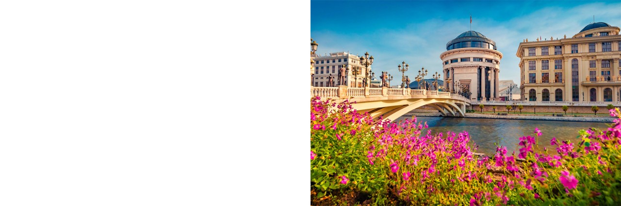 Das Bild zeigt eine über den Fluss verlaufende Brücke mit Straßenlaternen. Im Vordergrund sind pinke Blumen zu sehen. Auf der anderen Flussseite stehen drei markante beige Gebäude.