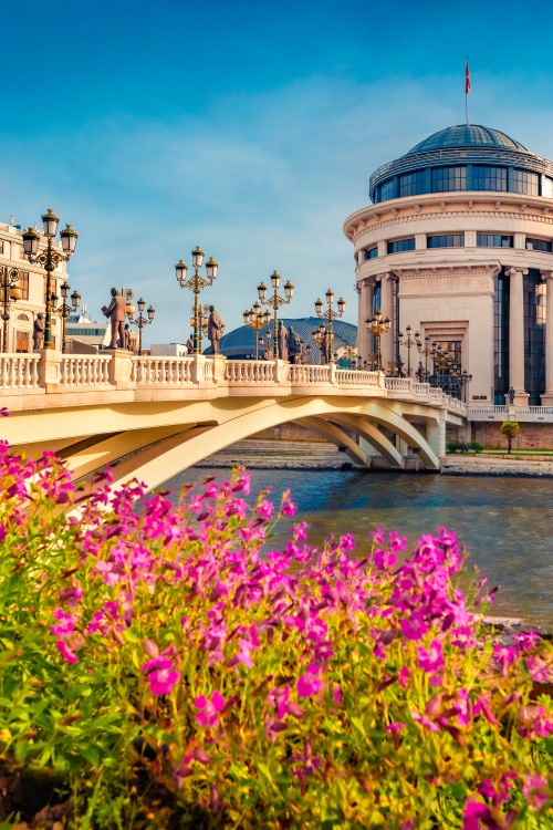 Das Bild zeigt eine über den Fluss verlaufende Brücke mit Straßenlaternen. Im Vordergrund sind pinke Blumen zu sehen. Auf der anderen Flussseite stehen drei markante beige Gebäude.