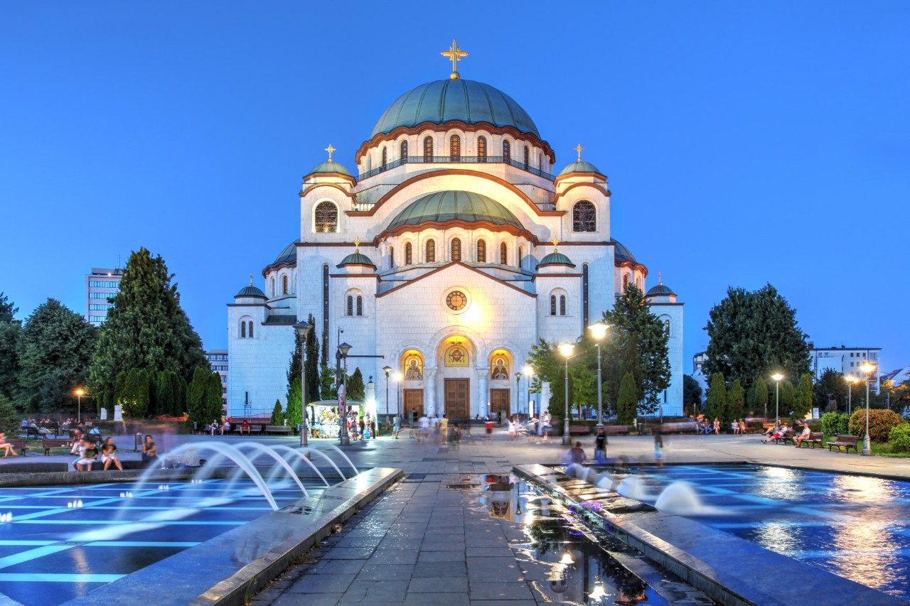 Frontalansicht einer großen orthodoxen Kathedrale mit einer mächtigen, türkisfarbenen Kuppel im Zentrum und mit einem goldenen Kreuz auf der Spitze im Abendlicht. Das Hauptschiff der weißen, beleuchteten Kathedrale ist mit mehreren kleineren Türmen gesäumt, drei bogenförmige Eingänge führen hinein. Auf dem Platz vor der Kirche rechts und links des Bildes sind zwei große, rechteckige, beleuchtete Springbrunnen zu sehen. Zu beiden Seiten der Kathedrale stehen Bäume.