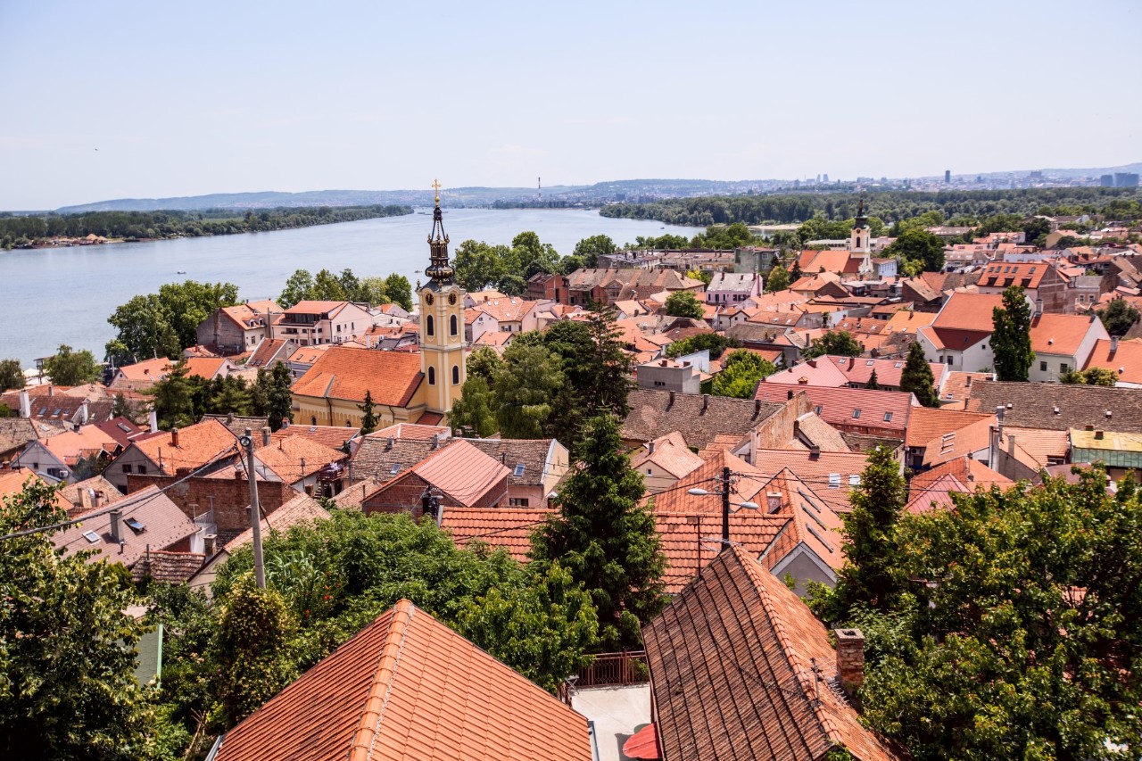 Blick von oben auf eine kleinere Stadt mit zahlreichen roten Ziegeldächern und einem Kirchturm im Bildzentrum. Im Hintergrund ist ein weiterer Kirchturm zu sehen, im linken Bildhintergrund ein Fluss.