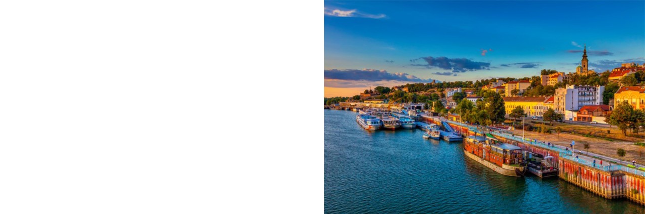 Blick von oben auf einen Hafen am Flussufer im Abendlicht. Im Hafen liegen mehrere Ausflugsboote. Das Flussufer ist mit Bäumen gesäumt, dahinter sind Häuser und in etwas weiterer Ferne ein Kirchturm einer Stadt zu sehen.