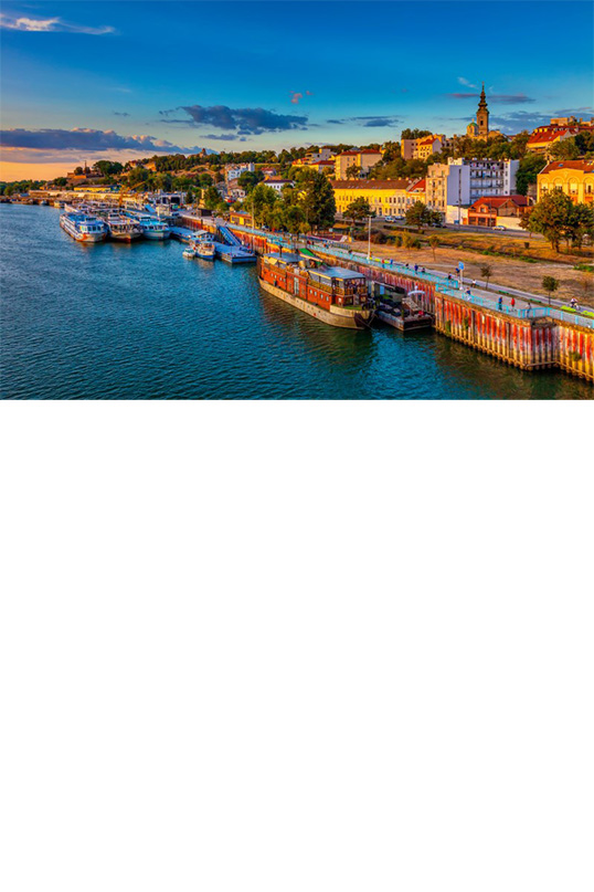 Blick von oben auf einen Hafen am Flussufer im Abendlicht. Im Hafen liegt ein Ausflugsboot. Das Flussufer ist mit Bäumen gesäumt, dahinter sind Häuser und in etwas weiterer Ferne ein Kirchturm einer Stadt zu sehen.