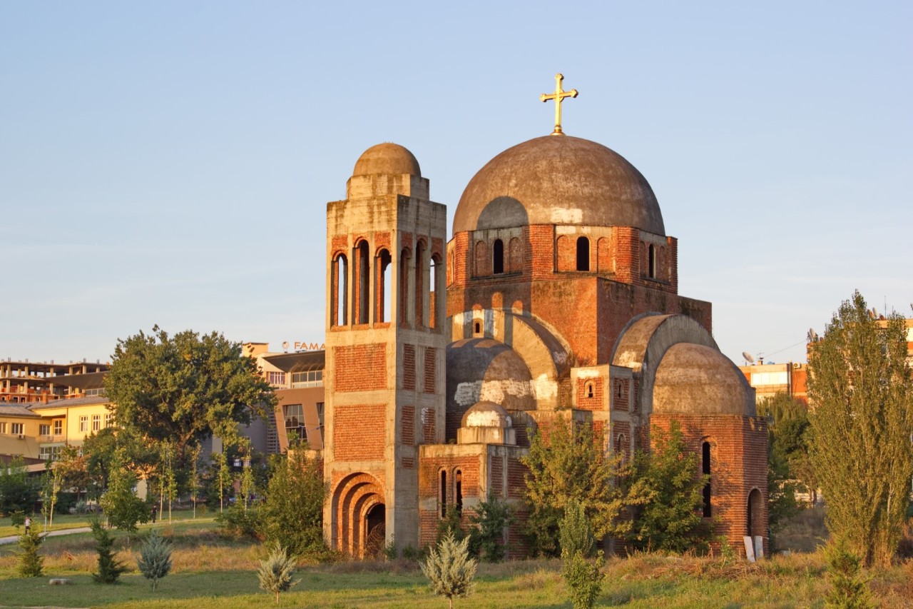 Zu sehen ist eine imposante Backsteinkirche mit einer großen, runden Hauptkuppel mit Kreuz und mehreren kleinen Kuppeln. Ein schmaler, viereckiger Turm mit Kuppel und offenen Bogenfenstern erhebt sich im Bildvordergrund. Rund um die Kirche sind grüne Rasenflächen, mehrere vereinzelte Bäume und Büsche sichtbar. Hinter der Kirche stehen Häuser.