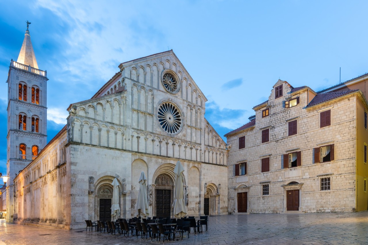 Blick auf eine große, helle Kirche mit einem rechteckigen Kirchenschiff und einem zweiteiligen Satteldach. Die Fassade wird von drei Eingängen und zwei übereinanderliegenden runden Rosenfenstern dominiert. Im Hintergrund ist ein eckiger Glockenturm mit Rundbogenfenstern und einem Spitzdach zu sehen. Vor der Kirche stehen auf einem großen Platz zahlreiche schwarze Stühle. Rechts neben der Kirche steht ein weiteres, dreistöckiges, helles Gebäude. Die Fenster sind mit dunkelroten Fensterläden verdeckt.