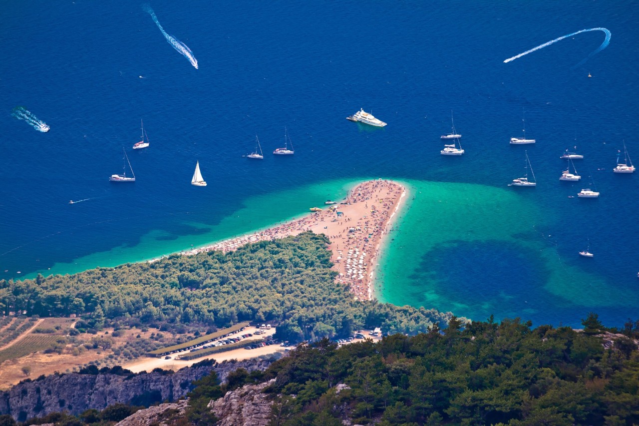 Blick von oben auf eine belebte Landzunge mit Strand, die wie ein Horn ins türkisblaue Meer ragt. Zahlreiche Boote liegen im tiefblauen Wasser. Umgeben ist der Strand von dichtem Wald.