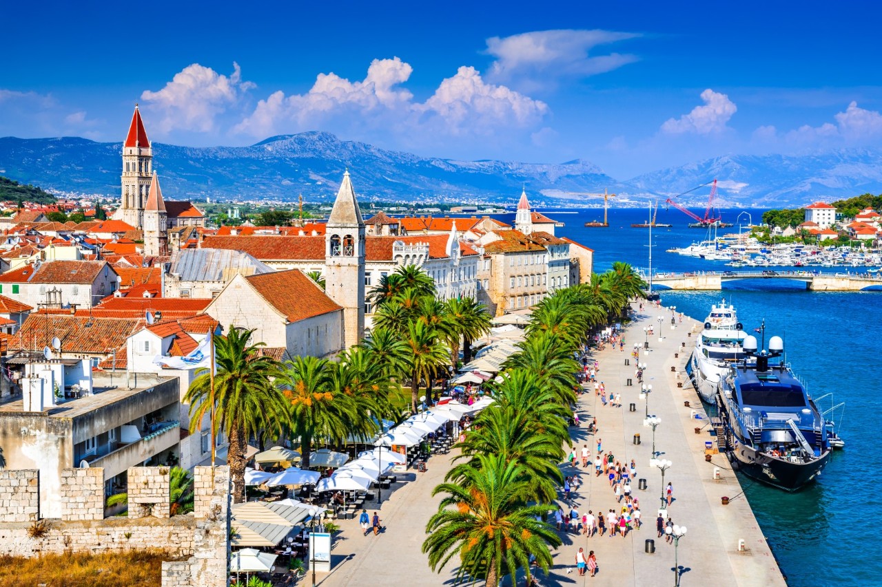 Das Bild zeigt einen Küstenort mit roten Dächern, historischen Gebäuden und Kirchtürmen. Entlang der palmengesäumten Uferpromenade gehen Menschen, während Yachten am Kai liegen. Im Hintergrund sind Berge und das Meer zu sehen.