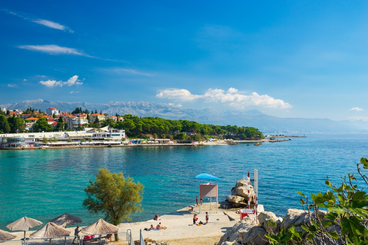 Das Bild zeigt eine Küstenlandschaft mit türkisblauem Meer, einem Strand mit Sonnenschirmen und Badenden im Vordergrund. Im Hintergrund ist eine grüne Landzunge mit Häusern zu sehen.