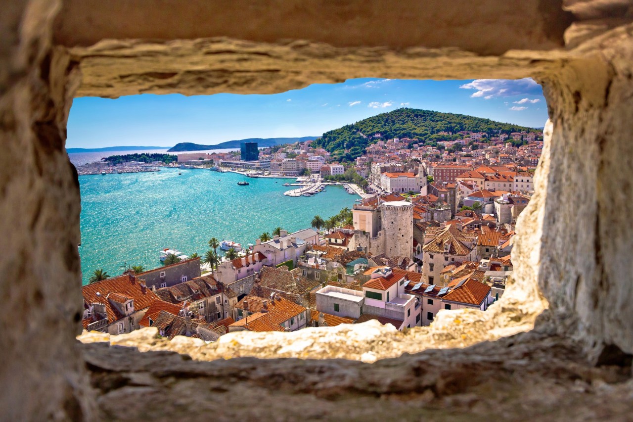Blick von oben durch eine steinerne Maueröffnung auf eine Stadt am Meer. Zu sehen sind rote Ziegeldächer, ein Hafen mit türkisblauem Wasser, Palmen entlang der Uferpromenade und ein bewaldeter Hügel im Hintergrund.