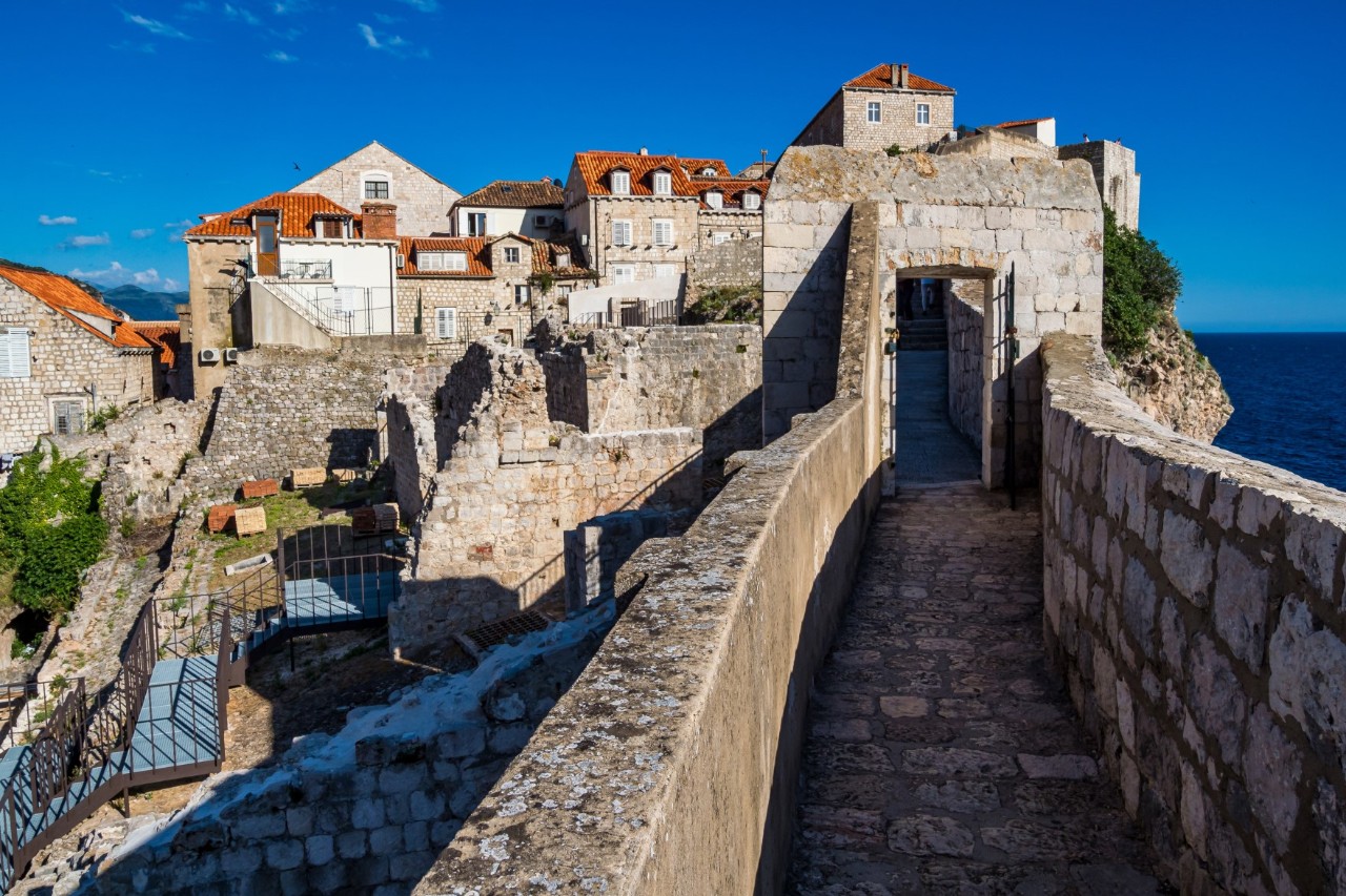 Blick auf eine begehbare Stadtmauer, die zu beiden Seiten von einer Mauer eingefasst ist. Im Bildhintergrund ist ein rechteckiges Tor innerhalb der Stadtmauer zu sehen, dahinter sind mehrere alte Häuser mit roten Ziegeldächern. Auf der linken Bildseite gibt es einige ruinenartige Mauerreste, durch die ein eingezäunter Weg führt. In der rechten hinteren Bildecke ist das Meer zu sehen.