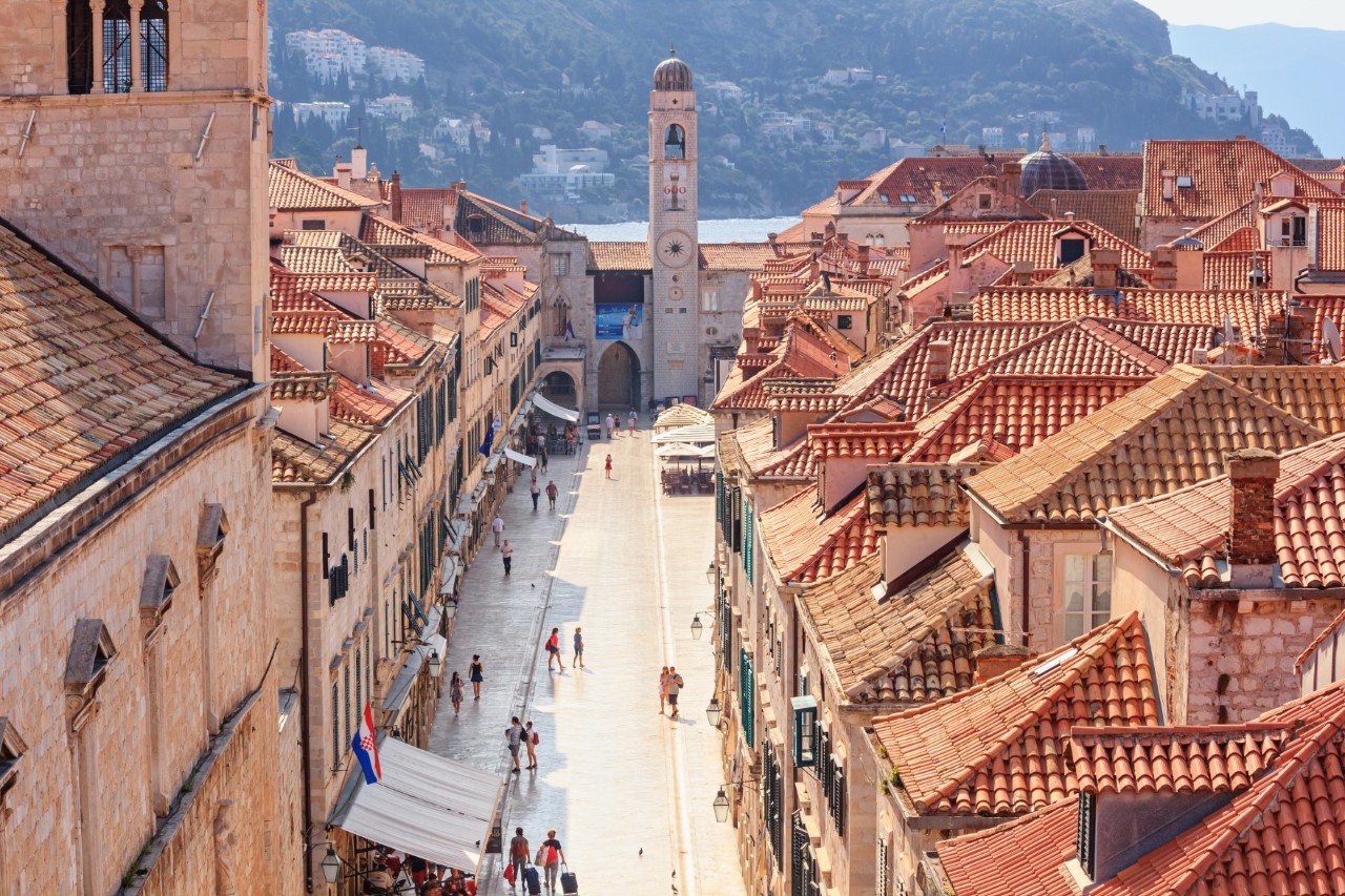 Blick von oben in die Fußgängerzone einer Altstadt, die auf einen schmalen rechteckigen Turm zuläuft. Auf der Straße sind zahlreiche Menschen unterwegs, auf der linken Bildseite ist eine Markise sichtbar. Die Gebäude zu beiden Seiten der Fußgängerzone haben markante, verwinkelte rote Ziegeldächer. Auf der linken Bildseite ragt ein höherer Gebäudeteil, vermutlich ein Turm, aus dem Bild. Im Bildhintergrund ist verschwommen eine felsige Küste zu sehen.