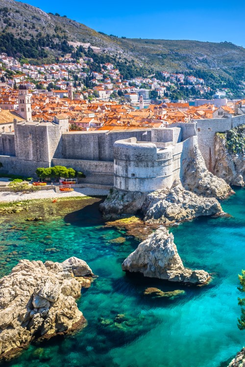 Ansicht einer Stadt am Meer, die auf eine Steilklippe gebaut und von einer massiven Stadtmauer umgeben ist. Zahlreiche rote Dächer sind zu sehen. Innerhalb der Stadtmauer befinden sich zwei größere Festungsanlagen. Aus dem türkisblauen Meer vor der Stadt ragen mehrere kleine Felsen empor. Auf der rechten Seite ragt eine grün bewachsene Klippe aus dem Bild.