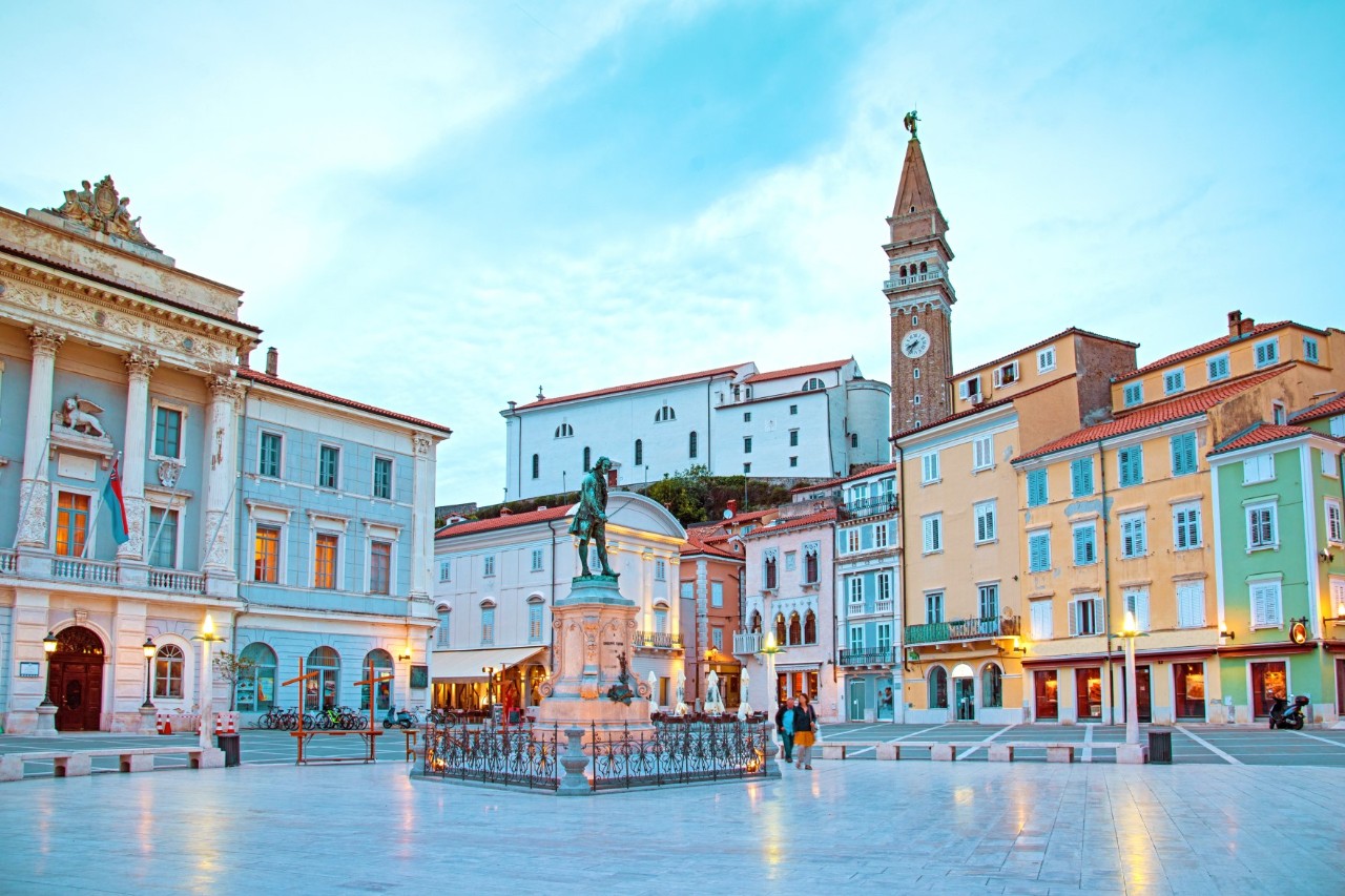 Das Bild zeigt einen zentralen Platz mit historischen Gebäuden in mediterraner Architektur. Eine Statue steht in der Mitte des Platzes. Im Hintergrund ragt ein Glockenturm empor, flankiert von pastellfarbenen Häusern und einer Kirche. 