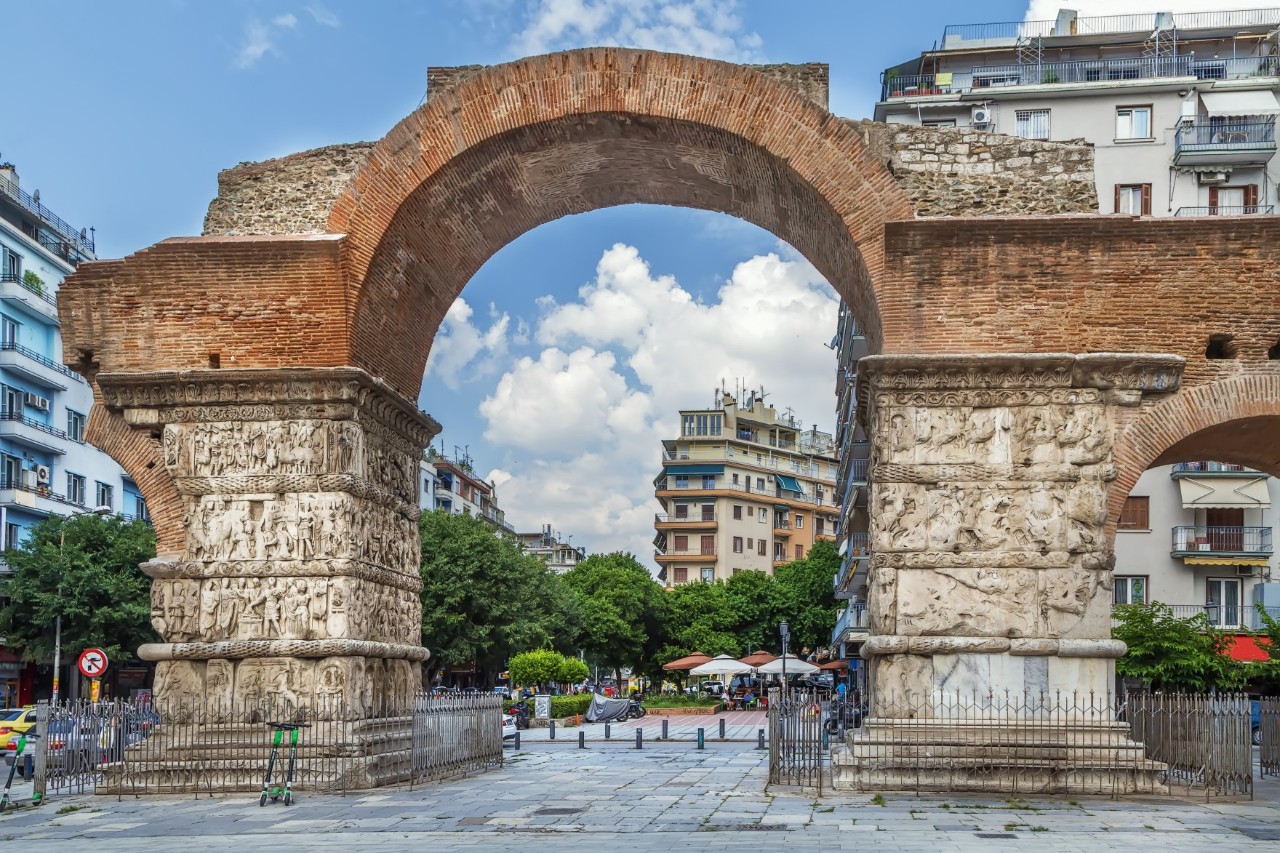 Die Aufnahme zeigt einen historischen Triumphbogen, der aus Ziegelsteinen und verzierten Steinsäulen besteht und mit Reliefs geschmückt ist. Er befindet sich in einer urbanen Umgebung mit modernen Gebäuden und Bäumen im Hintergrund.