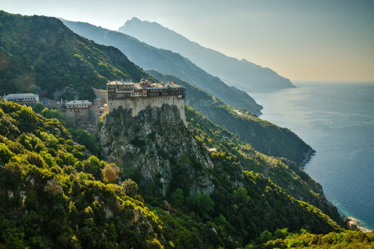 Die Aufnahme zeigt ein imposantes Kloster, das auf einem steilen Felsen hoch über dem Meer errichtet ist. Es ist von dicht bewaldeten Hügeln umgeben. Im Hintergrund und am rechten Bildrand erstreckt sich die Küstenlinie.