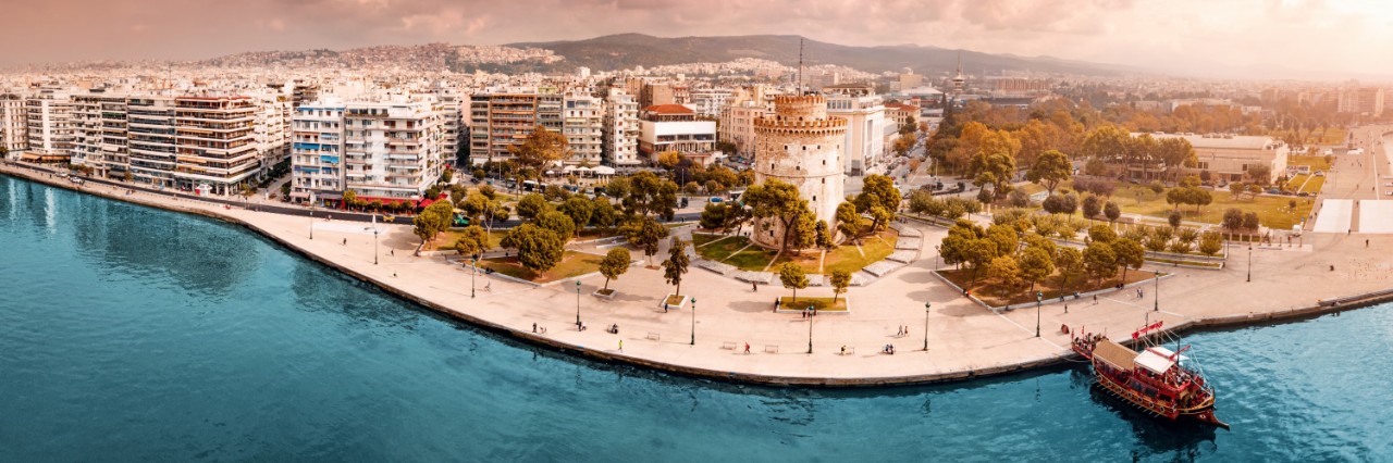 Das Bild zeigt eine beeindruckende Panoramaansicht der Uferpromenade von Thessaloniki. Im Zentrum steht der berühmte Weiße Turm, der umgeben ist von einem kleinen Park. Auf der linken Bildseite befinden sich moderne mehrstöckige Gebäude. Auf dem Wasser, das die untere Hälfte des Bildes dominiert, liegt ein traditionell aussehendes, rotes Holzboot. Im Hintergrund sind Hügel zu sehen.