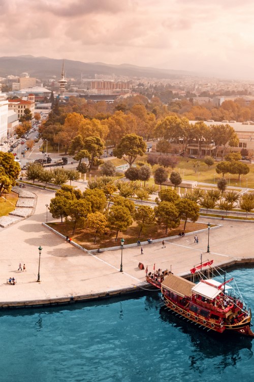 Das Bild zeigt eine beeindruckende Panoramaansicht der Uferpromenade von Thessaloniki. Im Zentrum steht der berühmte Weiße Turm, der umgeben ist von einem kleinen Park. Auf der linken Bildseite befinden sich moderne mehrstöckige Gebäude. Auf dem Wasser, das die untere Hälfte des Bildes dominiert, liegt ein traditionell aussehendes, rotes Holzboot. Im Hintergrund sind Hügel zu sehen.