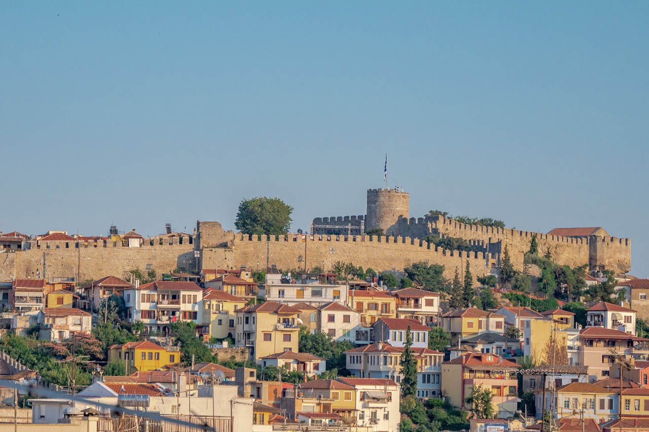 Historische Burganlage aus Stein auf einem Hügel. Die massiven Mauern und Türme stehen vor blauem Himmel, darunter breitet sich die Stadt Kavala aus.