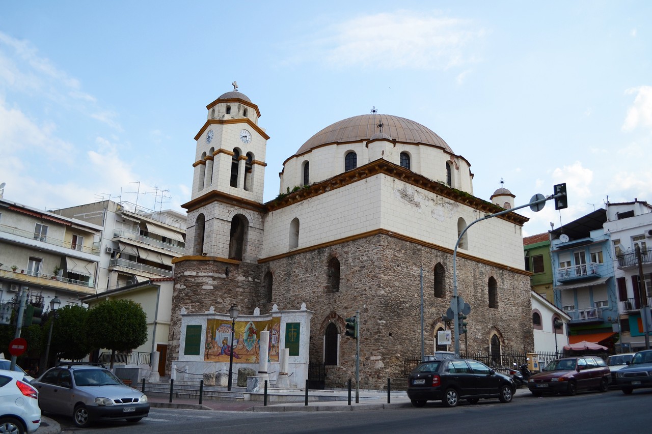 Außenansicht einer Steinkirche mit großem Runddach und schlankem Glockenturm in städtischer Umgebung. Im Vordergrund befindet sich ein kleiner Platz und Straßen auf denen Autos fahren. Im Hintergrund sind Gebäude und der blaue Himmel zu sehen.