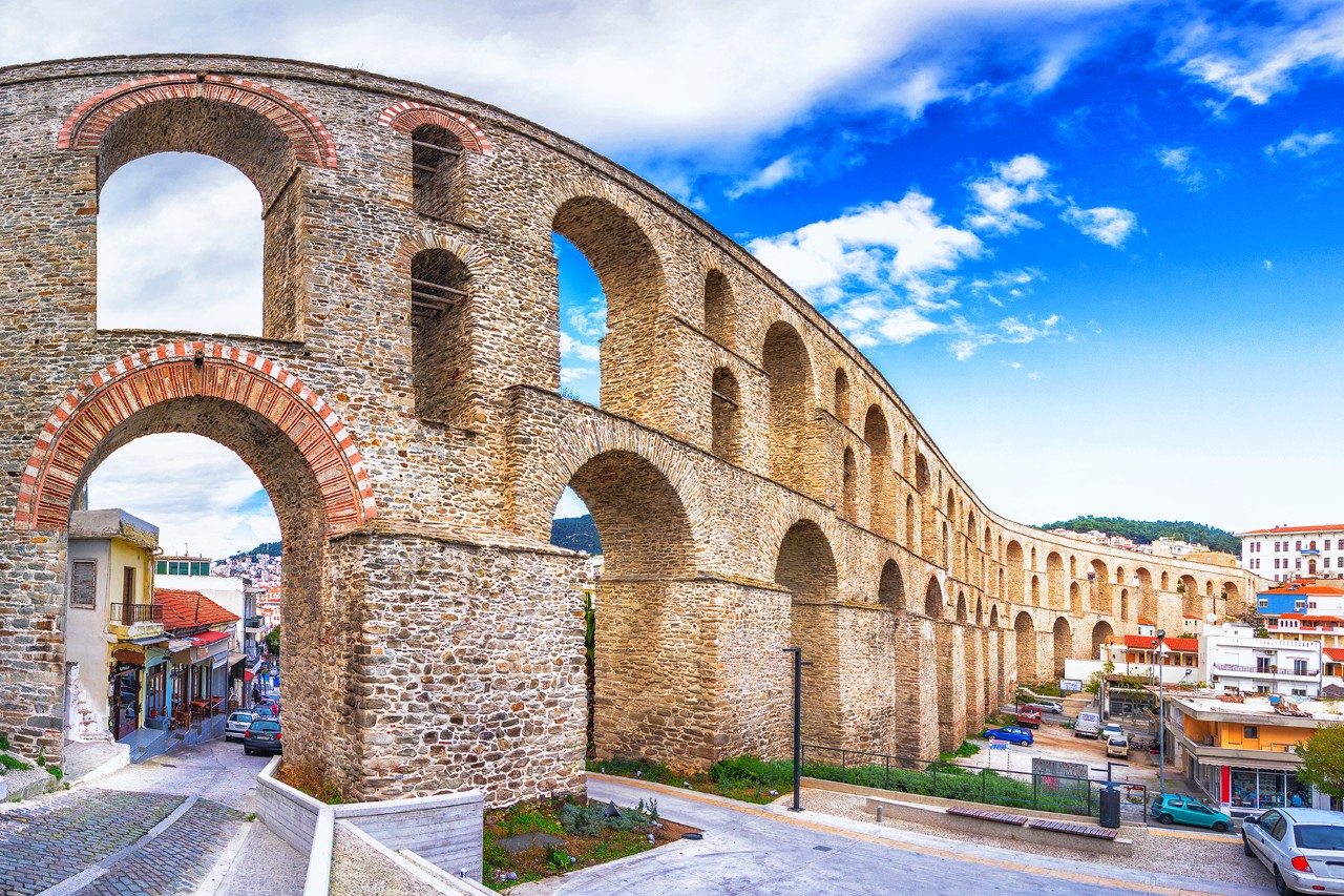 Großes historisches Aquädukt mit mehreren hohen Steinbögen, das sich durch das urbane Stadtgebiet von Kavala zieht. Im Vordergrund sind Straßen zu sehen, im Hintergrund Hügel sowie blauer Himmel mit Wolken.