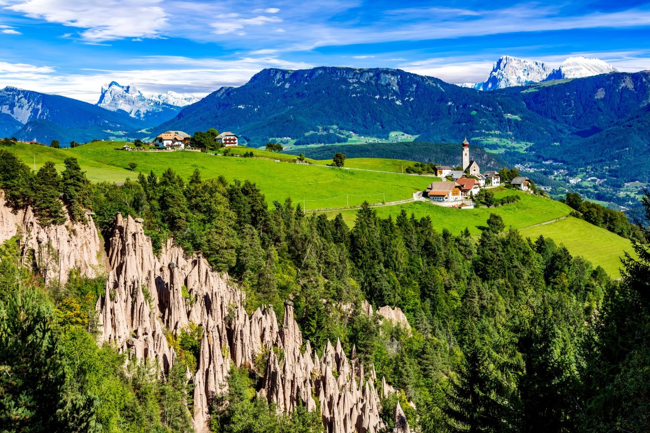 Spitze Felsformationen, die sich in einem Nadelwald erheben. Dahinter ist eine große, grüne Alm zu sehen mit zwei kleinen Bergdörfern, hinter denen sich Berge erheben.