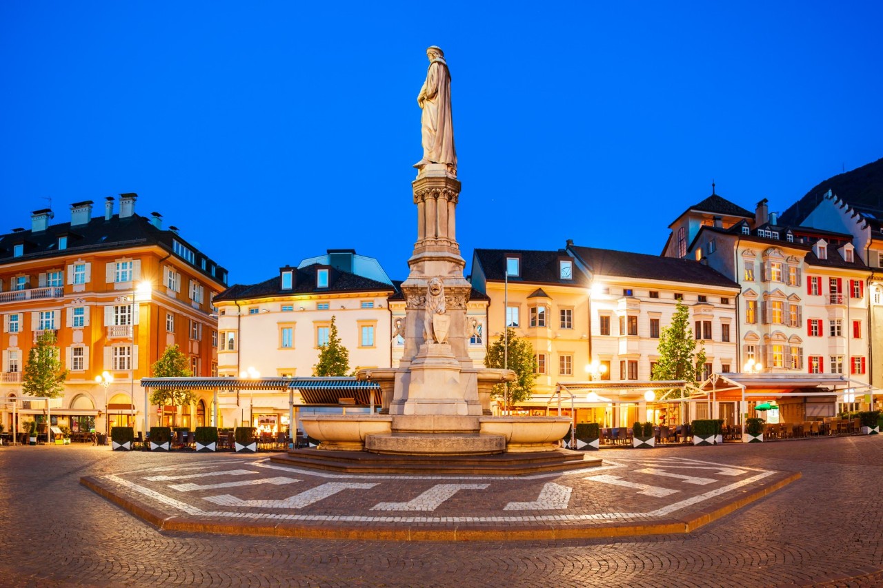 Großer Platz mit einer weißen Statue im Zentrum. Dahinter stehen historische, mehrstöckige Häuser, die in der Abenddämmerung erleuchtet sind.