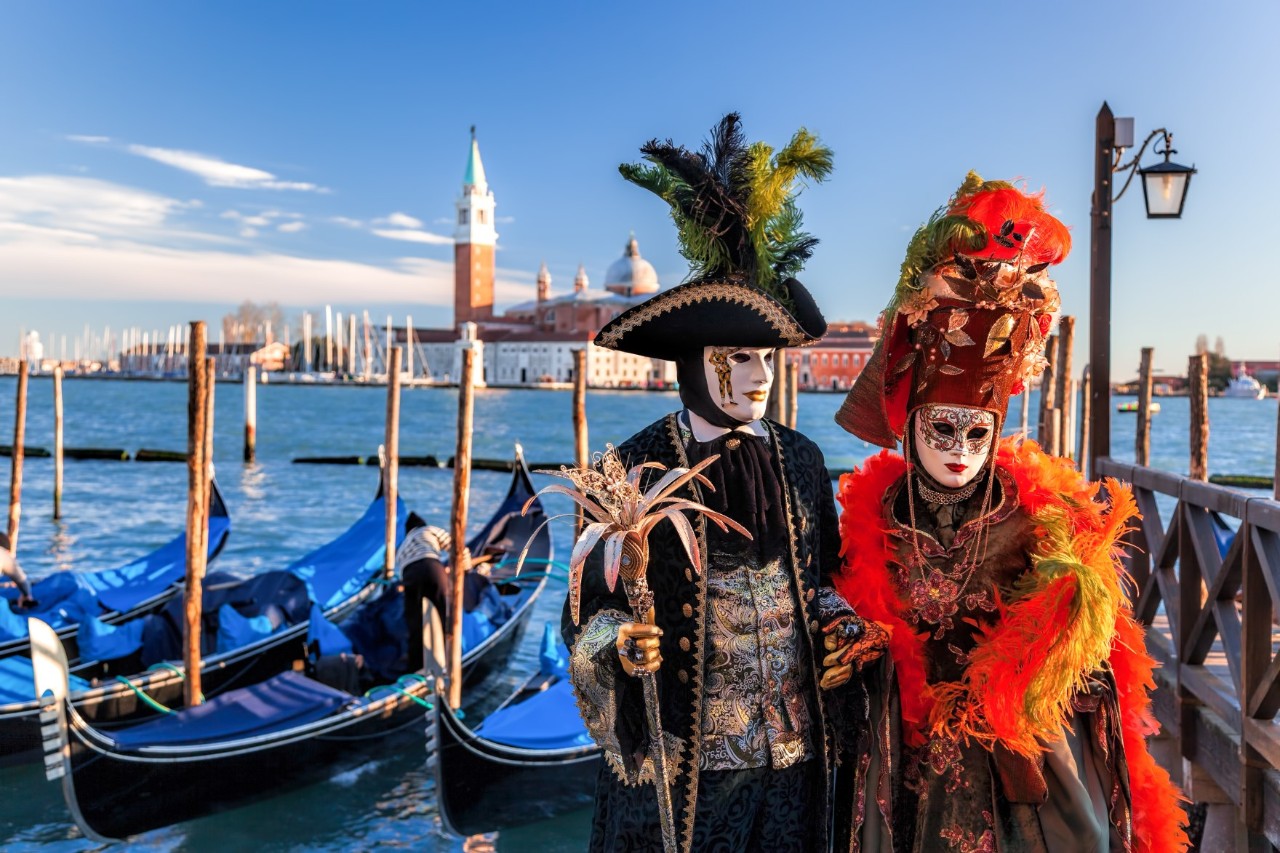 Paar in traditionellen venezianischen Karnevalskostümen, im Hintergrund festgemachte Gondeln, Masten, Insel und Kirche San Girgio-Maggiore. © Thomas Marek/stock.adobe.com 