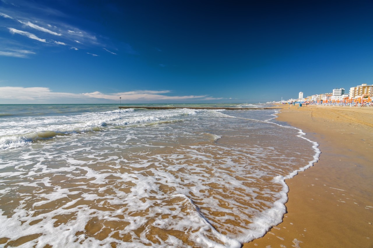 Links im Bild ist das Meer mit kleinen Wellen zu sehen. Rechts befindet sich der Sandstrand, im Hintergrund ist die Uferpromenade zu sehen. Der Himmel ist fast wolkenlos und erstrahlt in kräftigen Blautönen.