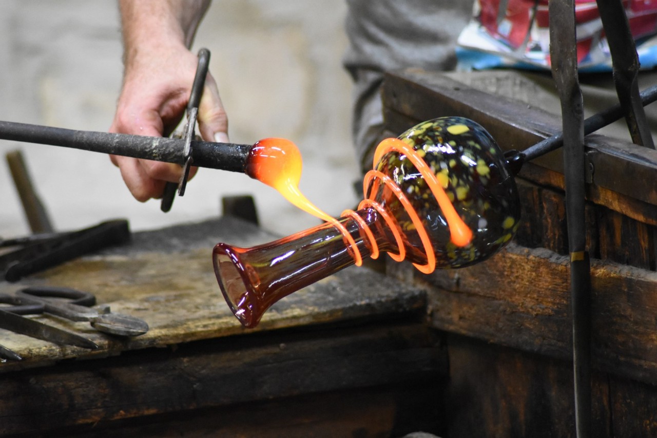Nahansicht des Vorgangs des Glasgießens. Heiße Glasfäden, die noch orangefarben leuchten, werden um eine kunstvolle Vase gewickelt werden.