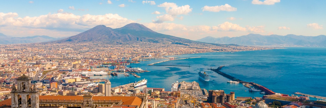 Luftansicht von Neapel, im Vordergrund sind die Altstadt und Häuser, rechts das Meer, am Horizont zeichnet sich der Vesuv ab, Himmel mit Cumulus-Wolken. © pfeifferv/stock.adobe.com