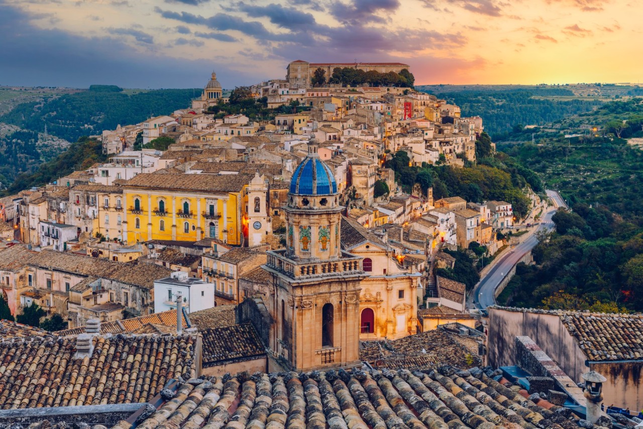 Die Häuser des auf einem Berg liegenden Ortes Ragusa bei Sonnenuntergang. Grüne Landschaften im Tal zu allen Seiten des Bergortes 