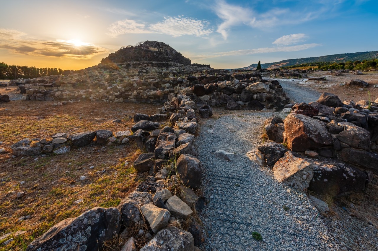 Das Bild zeigt eine archäologische Stätte mit einer antiken Nuraghe, einer charakteristischen Turmbauweise. Umgeben von steinernen Mauern und Ruinen liegt die Stätte im warmen Sonnenlicht.
