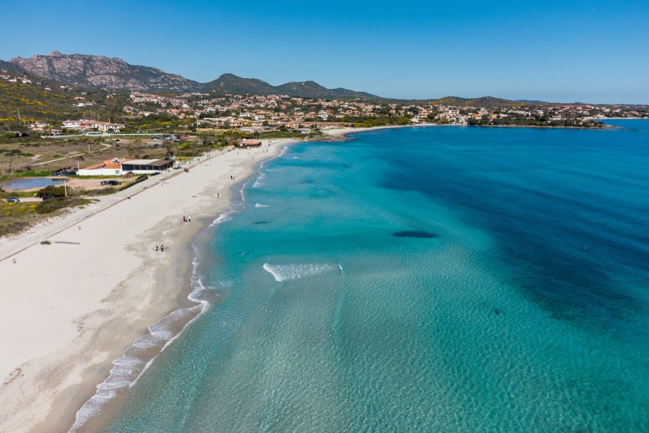 Das Bild zeigt einen langen, weißen Sandstrand, der von kristallklarem, türkisfarbenem Wasser gesäumt wird. Im Hintergrund erstrecken sich sanfte Hügel und eine Ortschaft mit Häusern, eingebettet in grüne Vegetation.