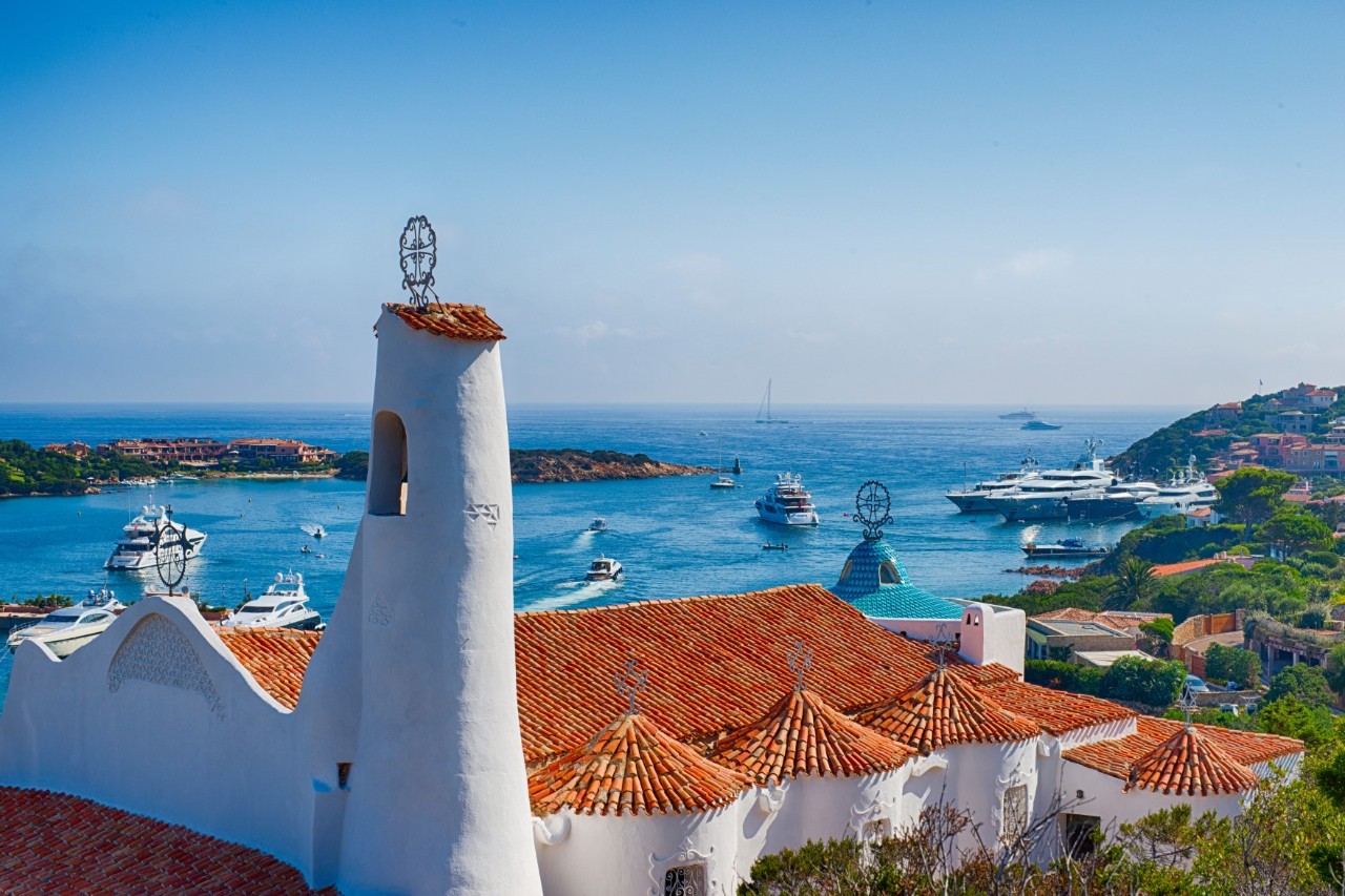 Das Bild zeigt eine mediterrane Küstenszene mit traditionellen Häusern, deren Dächer aus roten Ziegeln bestehen. Im Vordergrund ist ein markanter, schmaler weißer Turm zu sehen. Im Hintergrund erstreckt sich das blaue Meer. 