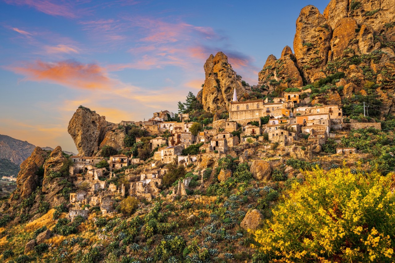 Das Bild zeigt ein Bergdorf in steiler Hanglage mit alten Steinhäusern, das sich in eine felsige Landschaft einfügt. Im Hintergrund ragen imposante Felsformationen empor. Im Vordergrund ist üppige grüne Vegetation sichtbar.