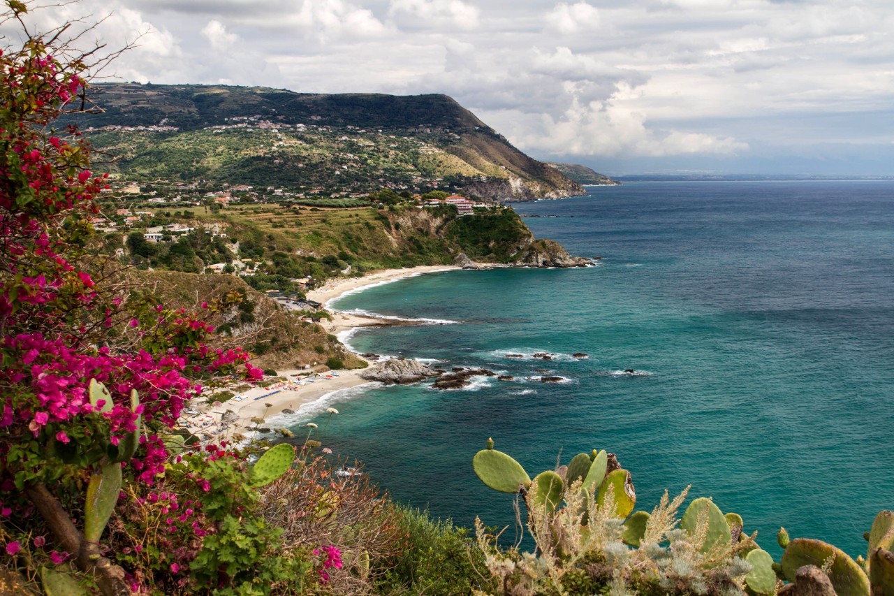 Das Bild zeigt eine Küstenlandschaft mit türkisblauem Meer, sandigen Buchten und felsigen Abschnitten. Im Vordergrund sind pinkfarbene Blumen und Kakteen, während im Hintergrund grüne Hügel und Häuser zu sehen sind.