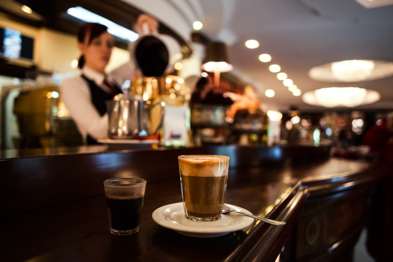 Kaffeehaus mit Frau an der Kaffeemaschine hinterm Tresen, beide unscharf im Hintergrund zu sehen, zwei kleine Gläser mit Kaffee im Vordergrund.