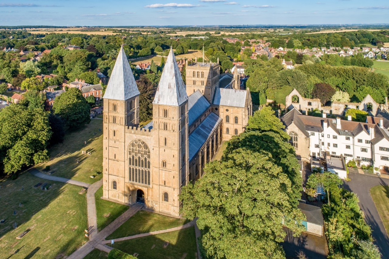 Blick aus der Vogelperspektive auf eine imposante beigefarbene Kathedrale mit zwei markanten Türmen und einem langen Kirchenschiff. Die Kirche ist von Grünflächen und Bäumen umgeben. Im Hintergrund verläuft eine ländliche Siedlung.