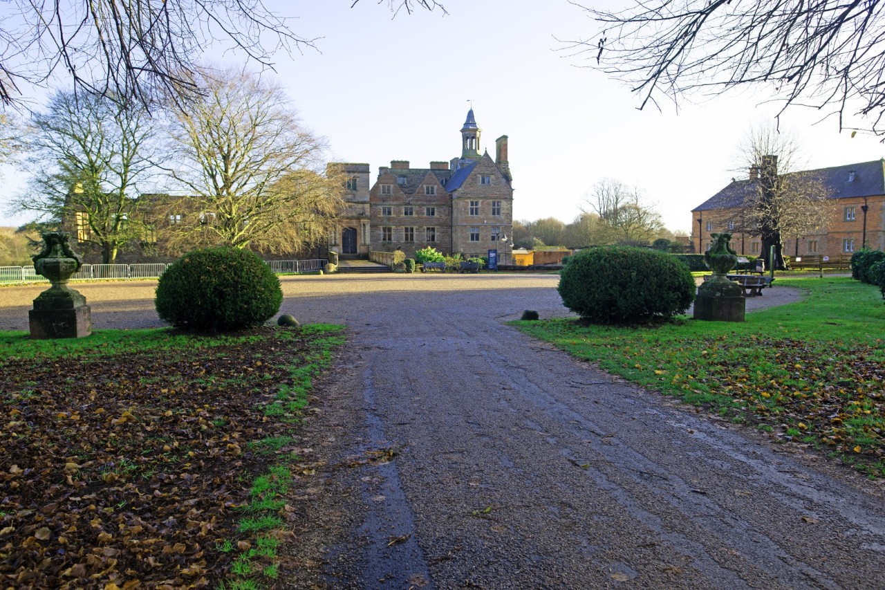 Das Bild zeigt eine historische Anlage mit einem großen Herrenhaus im Hintergrund. Ein Weg führt durch eine Gartenanlage, die von zwei dekorativen Steinskulpturen und symmetrisch platzierten Büschen gesäumt wird. Die Bäume sind teils kahl.