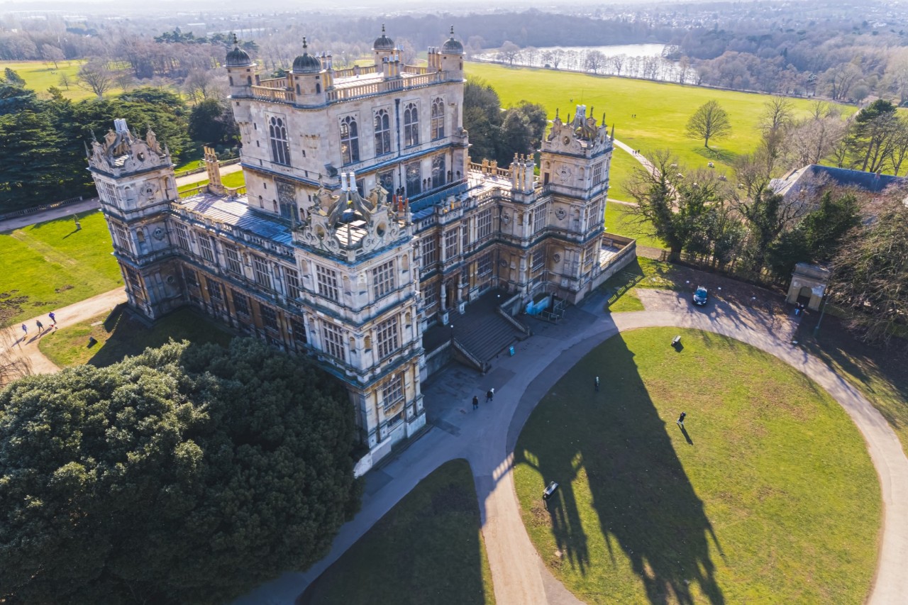 Das Bild zeigt ein altes Schloss aus der Vogelperspektive. Die Fassade ist aufwendig verziert, mit vielen Türmen und dekorativen Elementen. Das Gebäude liegt inmitten eines weitläufigen Parks mit grünen Wiesen und einem Fluss im Hintergrund.