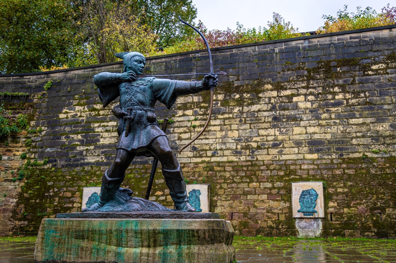 Das Bild zeigt eine bronzene Skulptur auf einem steinernen Sockel, die Robin Hood in einer dynamischen Pose darstellt. Er schießt mit gespanntem Bogen einen Pfeil ab. Die Statue steht vor einer alten dunkelgrau-beige-grünen Steinmauer. 