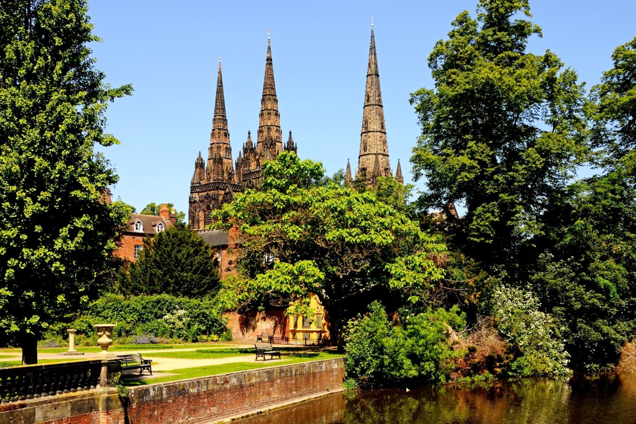 Das Bild zeigt eine gotische Kathedrale mit drei Spitztürmen, umgeben und teilweise verdeckt von Bäumen und gepflegten Grünflächen. Im Vordergrund verläuft ein kleiner Fluss entlang eines gepflegten Gartens mit Sitzbänken.