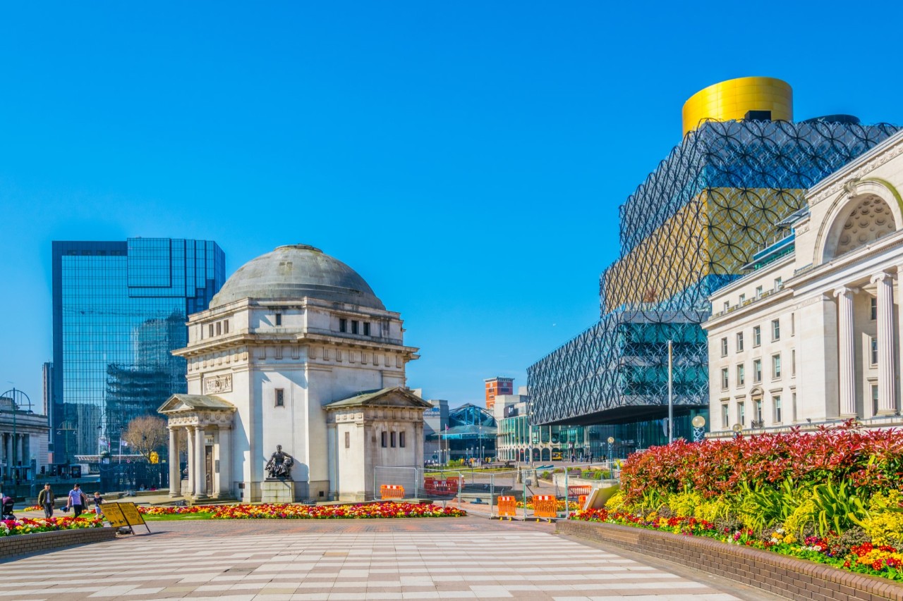 Das Bild zeigt einen menschenleeren Platz, umgeben von Blumenbeeten und mehreren historischen und modernen Gebäuden. Im Vordergrund steht ein rundes Denkmal mit einem kuppelförmigen Dach. Dahinter sind Wolkenkratzer mit Glasfassaden zu sehen. Rechts im Bild ist ein markantes, modernes Gebäude zu sehen, das durch seine auffällige Architektur mit einem Gittermuster und einer goldenen runden Kuppel hervorsticht. Der Himmel ist blau und klar. 