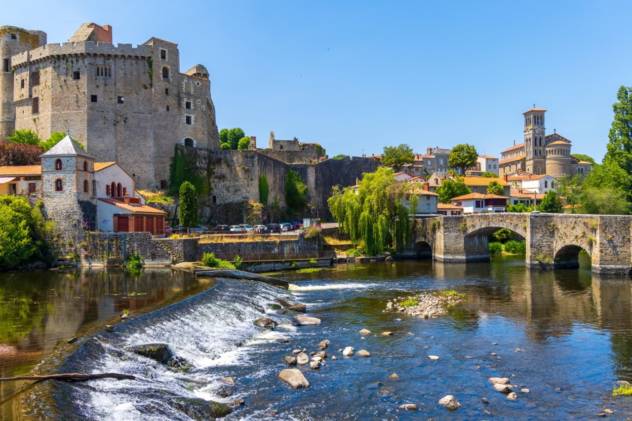 Das Bild zeigt links eine mittelalterliche Burg, die auf einer Anhöhe neben einem Fluss thront. Im Vordergrund fließt ein Fluss mit einer kleinen Kaskade über Steine. Rechts im Bild befindet sich eine alte Steinbrücke mit mehreren Bögen. Die Umgebung besteht aus kleineren Gebäuden und einer Kirche. Der Himmel ist blau und wolkenlos. 