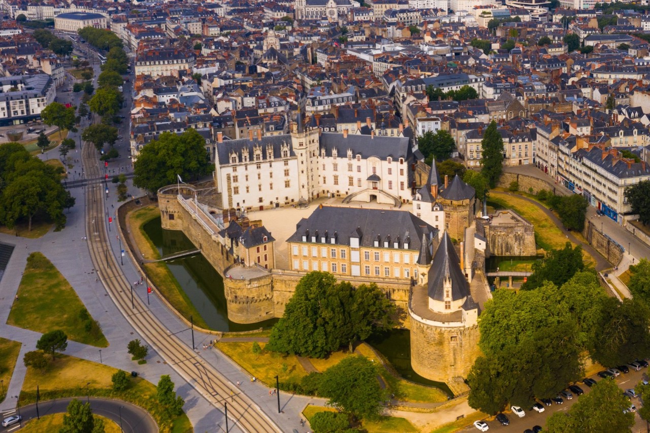 Luftaufnahme eines historischen Schlosses, umgeben von einer weitläufigen Stadt mit einem aus dem Häusermeer herausragenden Hochhaus. Links des Schlosses befindet sich eine menschenleere, breite Straße. Zum Schloss gehören eine Festungsmauer, Türme und ein Wassergraben, davor befinden sich Bäume und Grünflächen.