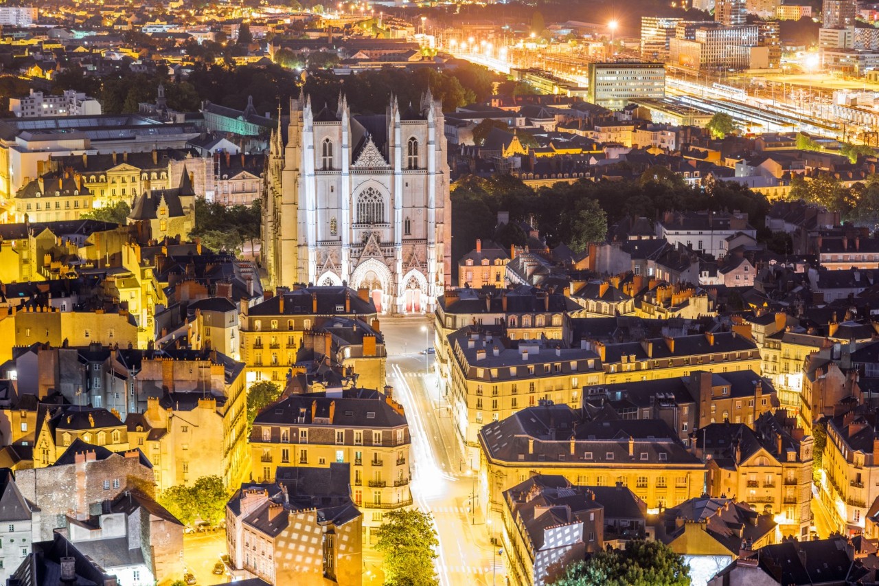 Das Bild zeigt eine nächtliche Stadtansicht mit einer beleuchteten Kathedrale im Zentrum. Die Umgebung besteht aus dicht stehenden Häusern, die in warmen Gelbtönen erleuchtet sind. 