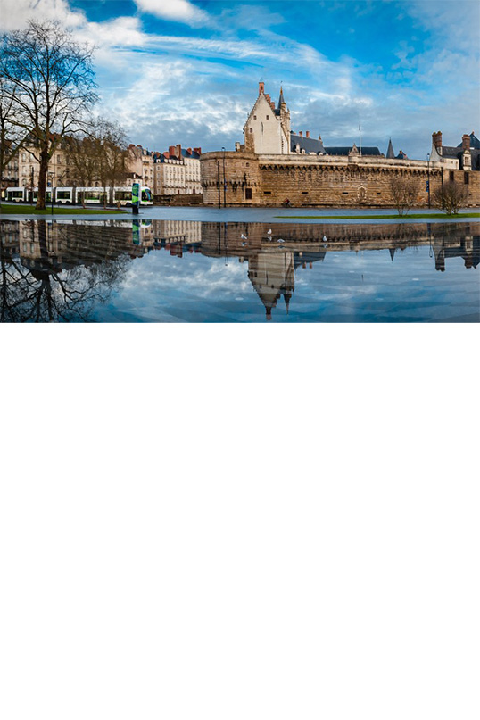 Das Bild zeigt eine Panoramaaufnahme eines Schlosses, das sich in einem ruhigen Wasserbecken spiegelt und von Häusern umgeben ist. Links ist eine Straßenbahn zu sehen. Die Bäume links und rechts im Bild sind kahl. Der Himmel ist teils von Schleierwolken durchzogen.