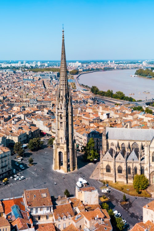 Luftbild von Bordeaux. Im Zentrum ist eine gotische Kathedrale mit einem langen Kirchenschiff sowie ein davorstehender, spitz zulaufender Turm zu sehen, der auf einen großen, freistehenden Platz gebaut ist. Rundum sieht man zahlreiche Häuser mit roten Dächern. Im Hintergrund ist ein Fluss zu sehen, über den eine Brücke führt. Am Horizont zeichnet sich ein blauer Himmelsstreifen ab.