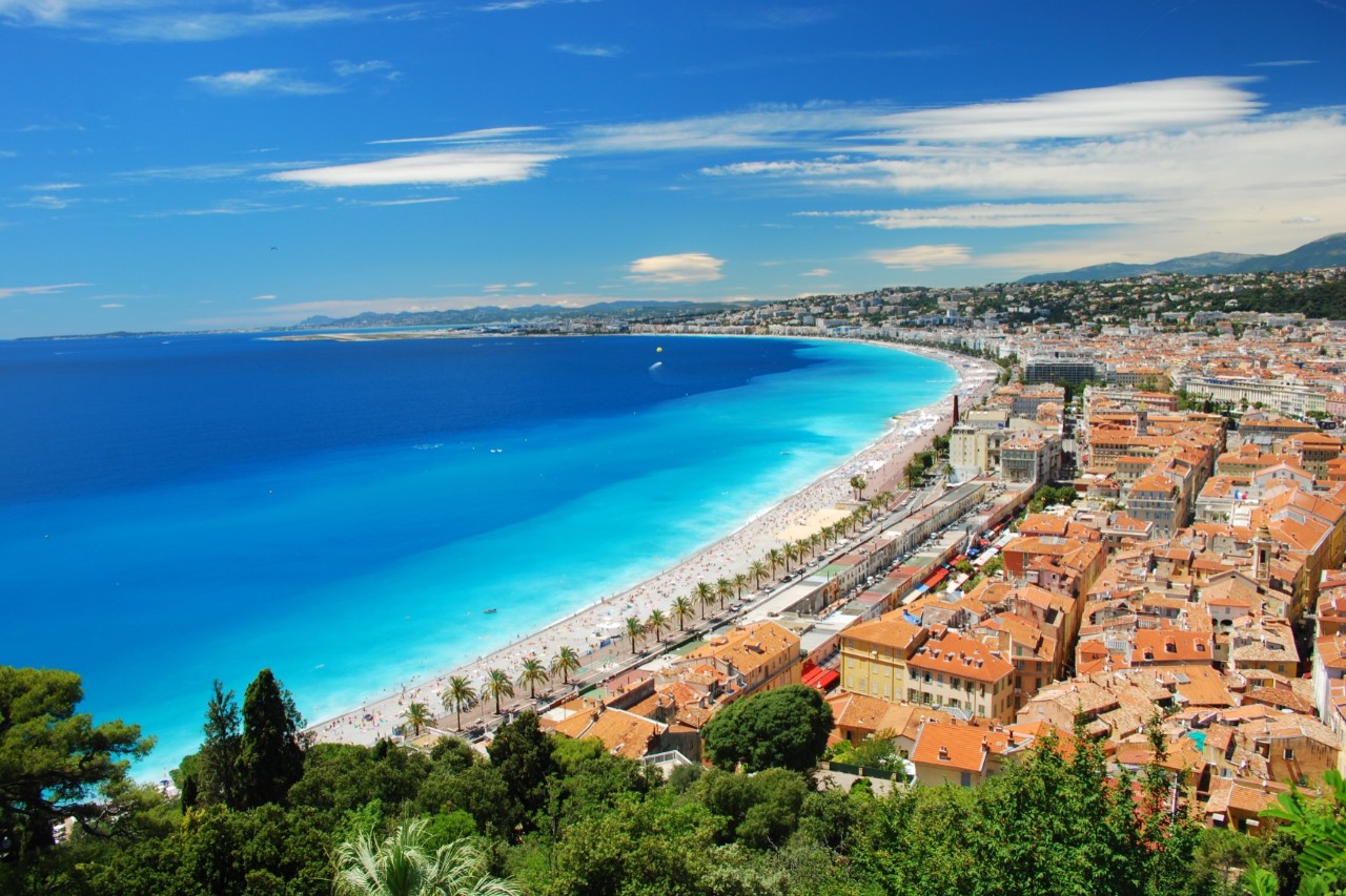 Vogelperspektive vom Park Colline du Château auf die Promenade des Anglais am Mittelmeer, davor die Altstadt und grüne Bäume im Park, türkisblaues Meer 