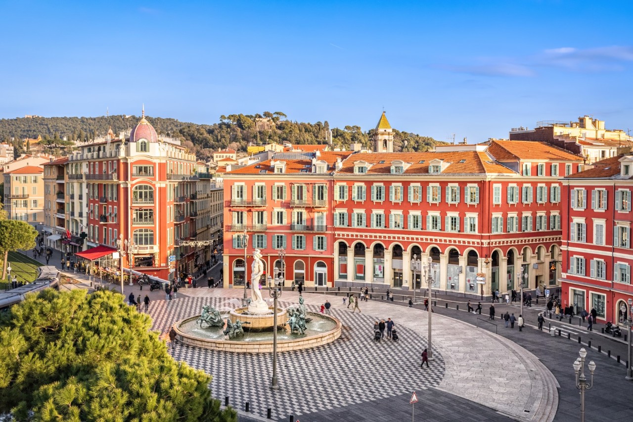 Place Masséna mit roten Häusern und weißen Fenstern, vereinzelte Menschengruppen, kleinen Läden und Springbrunnen mit Skulptur 