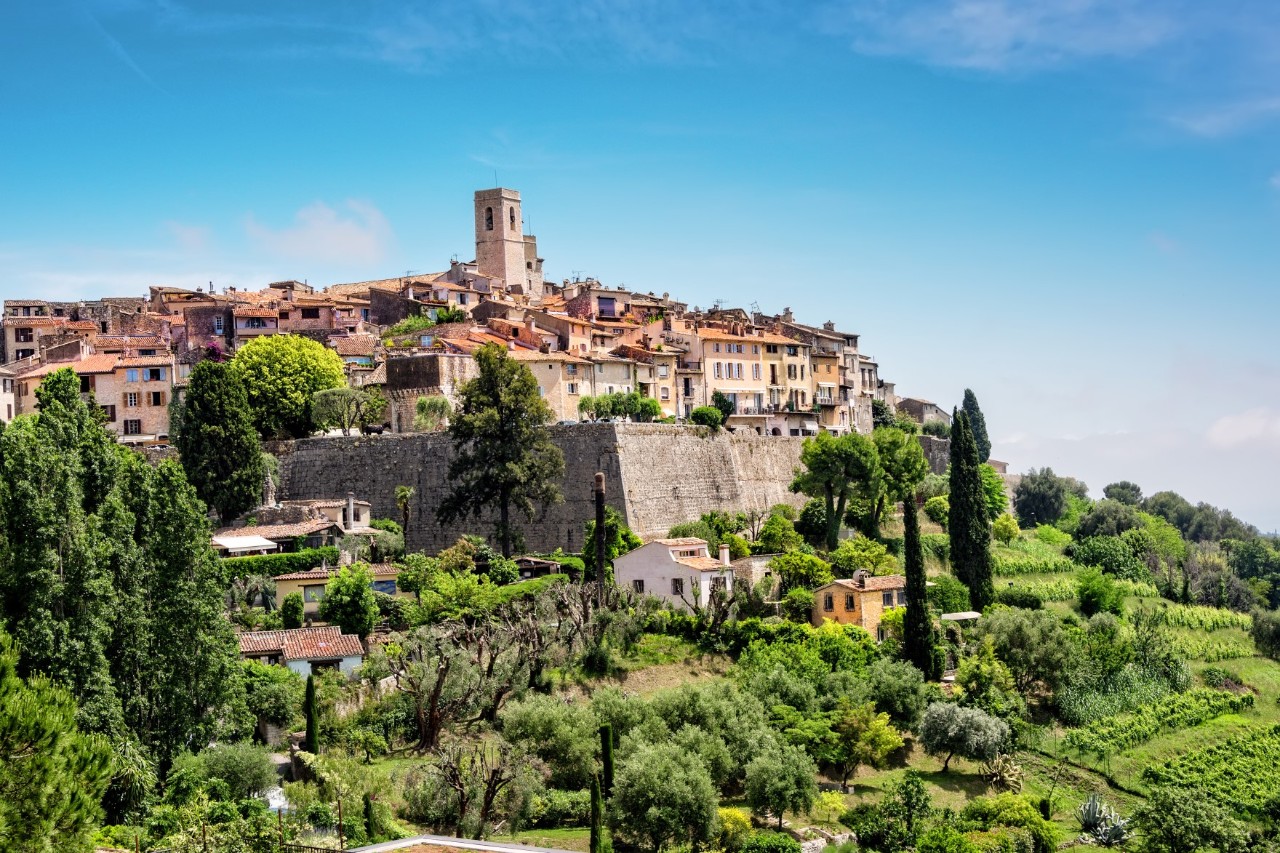 Saint-Paul-de-Vence auf einem Hügel mit Stadtmauer und Kirchturm, im Vordergrund kleinere Häuser im Grünen 